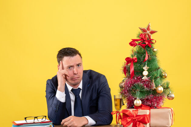 行政人员抑郁的男人坐在圣诞树旁的桌子前 黄色的礼物男性会议沮丧的男人