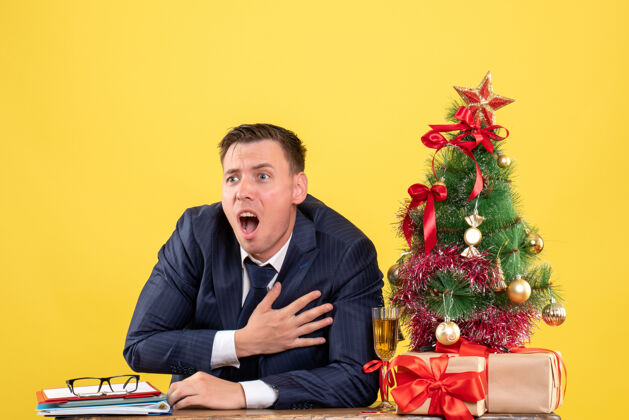 激动前视图焦躁不安的年轻人抱着胸脯坐在圣诞树旁的桌子旁 黄色的墙上摆着礼物圣诞节商务男人