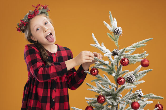 装饰戴着圣诞花圈的小女孩穿着格子裙装饰圣诞树快乐快乐地伸出舌头在橙色的背景上橙色快乐树