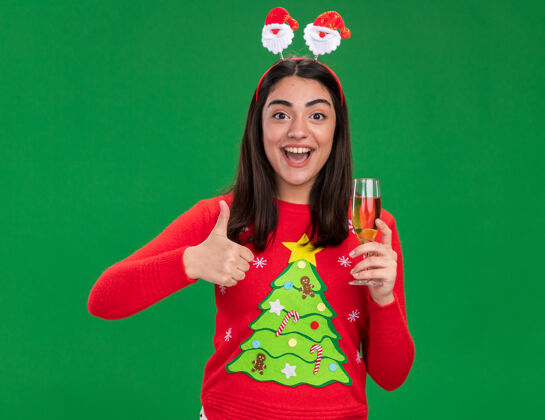 圣诞老人快乐的年轻白人女孩 戴着圣诞老人的头巾 竖起大拇指 拿着一杯香槟 背景是绿色的 有复制空间香槟女孩举行