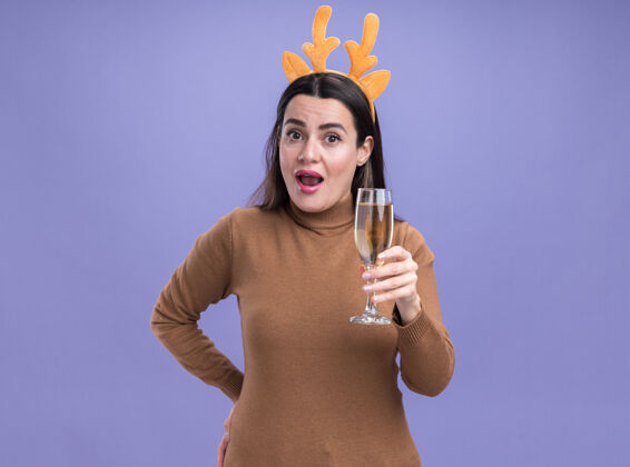 呼啦圈惊讶的年轻漂亮女孩穿着棕色毛衣 戴着圣诞发箍 拿着一杯香槟 背景是蓝色的毛衣头发女孩