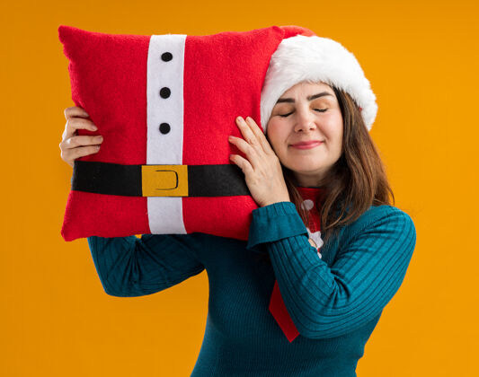 帽子高兴的成年白人妇女与圣诞帽和圣诞领带举行装饰枕头孤立的橙色背景与复制空间圣诞老人枕头装饰