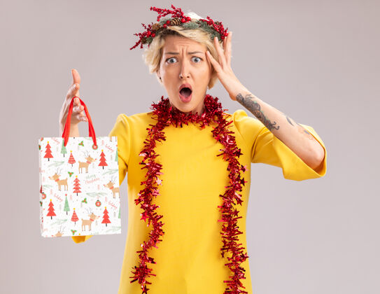 脖子关心的金发女郎头戴圣诞花环 脖子上戴着金箔花环 手里拿着圣诞礼品袋 看起来像是在白墙上孤立的头周围圣诞节花环