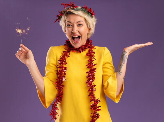 圣诞节令人印象深刻的年轻金发女子头戴圣诞花环 脖子上戴着金箔花环 手里拿着节日火花灯 在紫色的墙上孤立着一只空手空花环闪闪发光