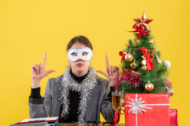 鸡尾酒正面图：戴着面具的年轻女孩坐在桌旁 手指拿着东西 圣诞树和鸡尾酒礼物成人女孩表演者