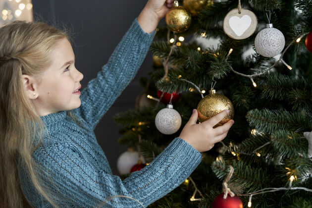 室内可爱的女孩把装饰品挂在圣诞树上传统准备快乐