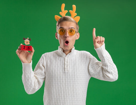 鹿角给人印象深刻的年轻帅哥戴着驯鹿鹿角头带戴着眼镜拿着圣诞树玩具和日期指着隔离在绿色墙上印象深刻穿着指向