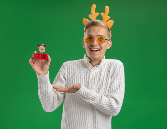 驯鹿令人印象深刻的年轻帅哥戴着驯鹿鹿角头带戴着眼镜拿着圣诞树玩具和日期指着它与手隔离在绿色墙上的复制空间印象深刻穿着帅气