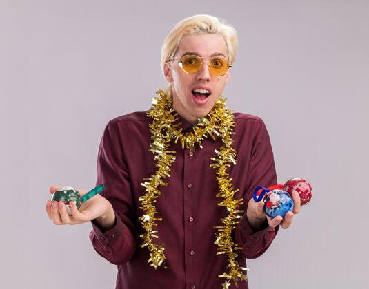 花环令人印象深刻的年轻金发男子戴着眼镜 脖子上戴着金属丝花环 手里拿着圣诞饰品 看着隔离在白色背景上的相机金发饰品周围