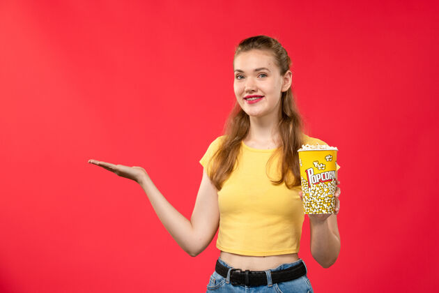 年轻的正面图年轻女性在电影院里拿着爆米花 微笑着站在红墙电影院里看女性欢乐时光电影爆米花电影女的