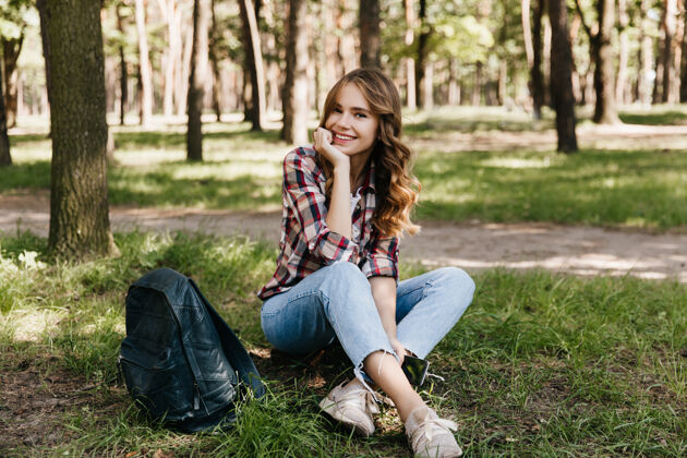 休息迷人的女游客带着背包在公园里摆姿势户外拍摄的迷人卷发女孩微笑着坐在草地上金发美丽背包客