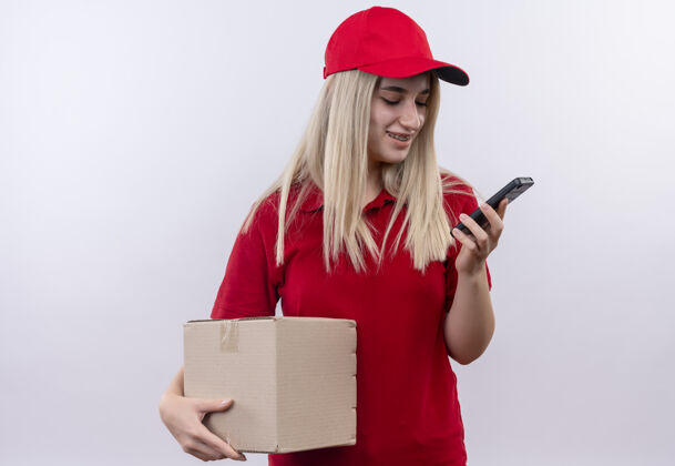 女孩微笑的小女孩穿着红色t恤 戴着帽子 戴着牙套 拿着盒子 在孤立的白色背景下看着手上的手机手送货牙科