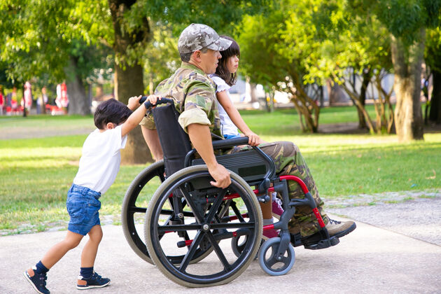 军队残疾退役军人带着孩子在公园散步女孩坐在爸爸的腿上 男孩推着轮椅退伍军人或残疾概念高加索制服退伍军人