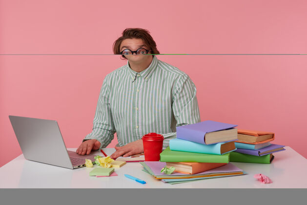 书年轻的黑发男性穿着条纹衬衫戴着眼镜在室内摆姿势 坐在那里拿着很多书和现代笔记本电脑 皱着额头扬起眉毛笔记本漂亮男人