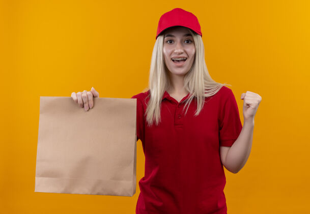 年轻微笑的小女孩穿着红色t恤 戴着帽子 戴着牙套 拿着纸袋 在孤立的橙色背景上显示“是”的手势红色牙科送货