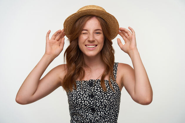 自然摄影棚拍摄了一位快乐美丽的年轻女士 红发卷曲 站在白色的背景上 举手把船夫的帽子戴在头上 对着镜头眨眼 积极地微笑着漂亮手欢快