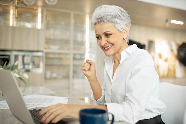 女性成功的成熟女作家坐在办公桌前 手里拿着手提电脑 文件和咖啡 脸上洋溢着幸福的表情 因为她按时完成了工作成功成功办公室