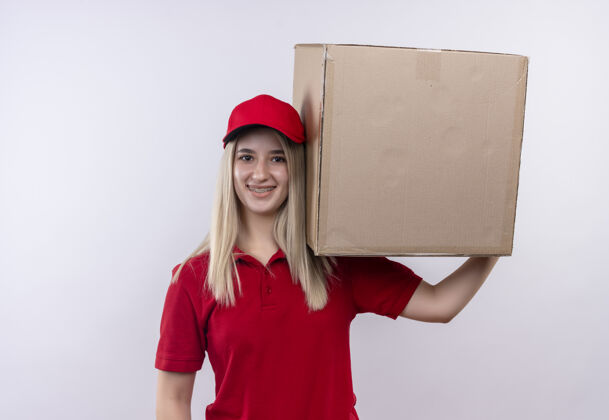 女孩微笑的小女孩穿着红色t恤 戴着帽子 戴着牙套 肩上扛着一个大盒子 背景是白色的白色肩膀牙科