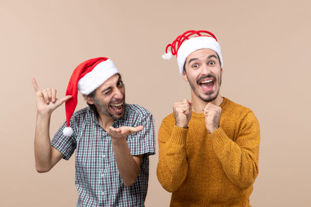 冲压正面图两个戴着圣诞帽的快乐的家伙一个眨着眼睛 另一个在米色孤立的背景上用拳头伙计们成人眨眼