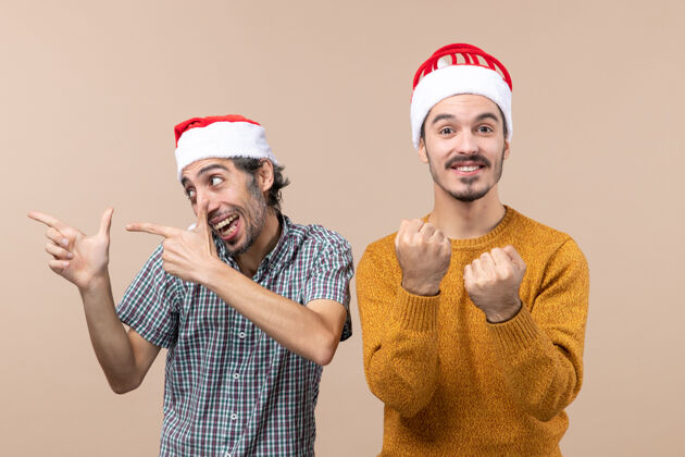 圣诞老人正面图两个戴着圣诞帽的惊讶的家伙 一个看着左边 另一个站在米色孤立的背景上打拳两个惊讶的家伙看伙计们