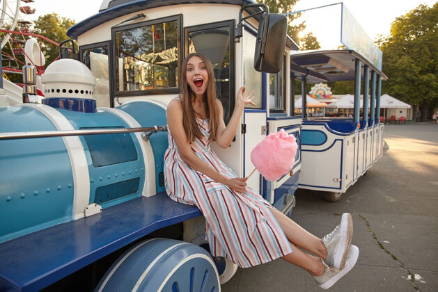 快乐在温暖的夏日 一位穿着浅色长裙的快乐迷人的女士坐在游乐园的蒸汽火车上 张大嘴巴 手里拿着粉色的棉花糖摆姿势棕色快乐公园