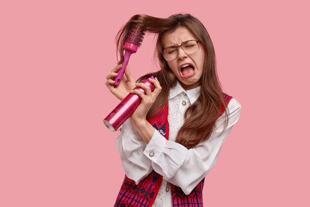 美发师惊慌失措的女人头发有问题 不能刷牙 面部表情低落 拿着发刷和发胶 约会迟到 穿着旧时髦的衣服 隔离在粉红色的墙上女人发胶摆姿势