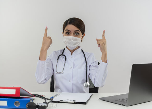 惊讶惊讶的年轻女医生穿着医用长袍 戴着听诊器 戴着医用口罩坐在办公桌上 用医用工具指着电脑上的复印空间穿女工作