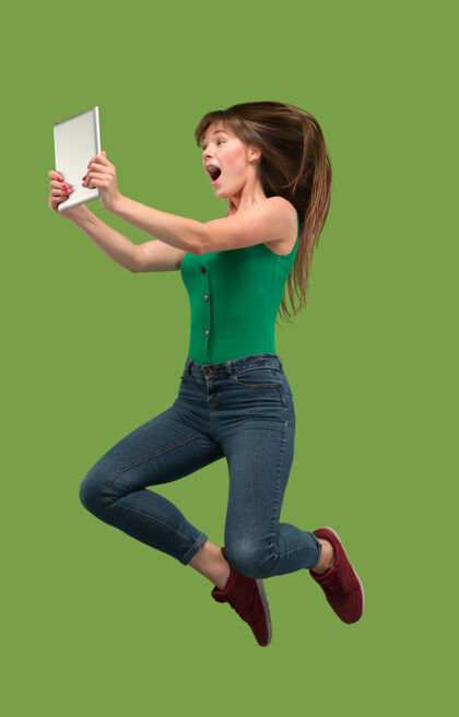 能源使用笔记本电脑或平板电脑小玩意在绿色工作室背景上跳跃的年轻女子在运动或运动中奔跑的女孩人类情感和面部表情概念现代生活中的小玩意携带休闲持有
