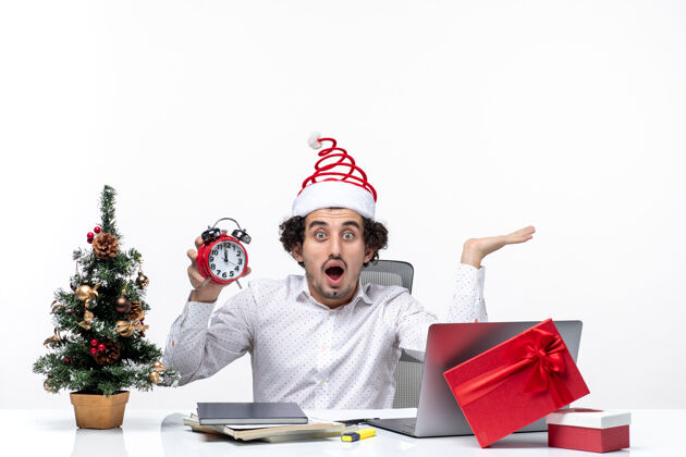 办公室年轻的商务人士戴着圣诞老人的帽子 展示着时钟 坐在白色背景的办公室里圣诞老人时钟工作
