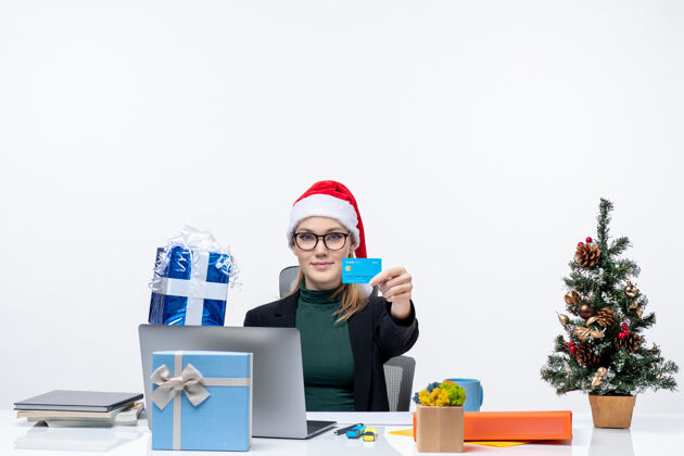 商务女人自信的女商人戴着圣诞老人的帽子 戴着眼镜坐在桌子旁 手里拿着圣诞礼物和白色背景的银行卡电脑银行抱着