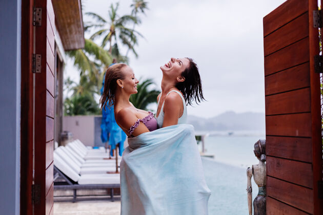 生活方式雨天 两个穿着比基尼的快乐女人在别墅外游泳池边用沙滩巾度假的户外写真饮料微笑放松