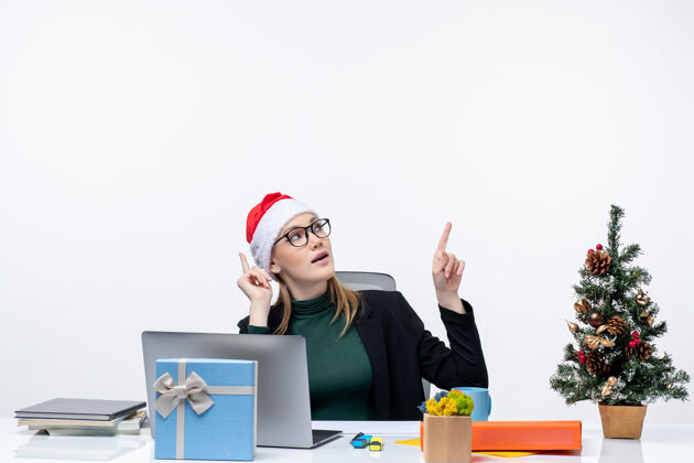 圣诞树一位自信的年轻女士 戴着圣诞老人帽 坐在一张桌子旁 桌子上放着圣诞树和一份礼物 在白色背景的左侧指向上方笔记本左边笔记本