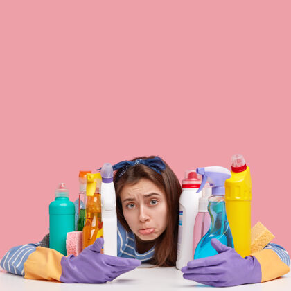 房间清洁概念心烦意乱的女人把下唇包起来 戴上防护手套 在带清洁产品的彩色瓶子旁摆姿势家庭面部家庭