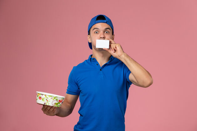 男人正面图身穿蓝色制服披肩的年轻男性信使手持白色卡片和浅粉色墙上的圆形投递碗男性递送卡片
