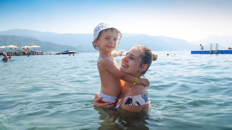 玩耍微笑的年輕母親站在海里抱著她蹣跚學步的小兒子的畫像自然嬰兒擁抱