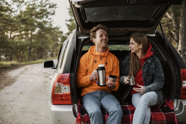男人笑脸情侣在车后备箱里享受热饮料饮料探险旅行