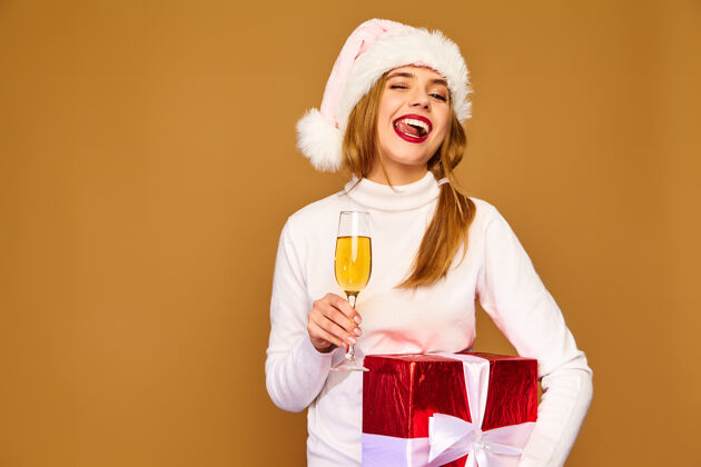 年轻戴着圣诞帽和大礼盒的模特在金墙上喝香槟时尚眨眼庆祝