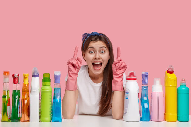 清洁剂乐观的年轻女佣用食指向上指 戴着头带和橡胶手套 在天花板上展示一些令人敬畏的东西瓶子T恤乐观
