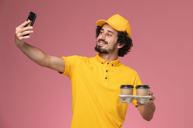 工作正面图身着黄色制服的男信使手持棕色的咖啡杯在粉色墙上拍照拿走粉色人