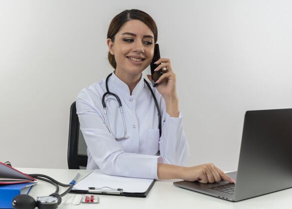 电脑微笑的年轻女医生穿着医用长袍 带听诊器 坐在办公桌旁 用电脑和医疗工具 用电话和笔记本电脑 还有复印空间用过的戴着医生