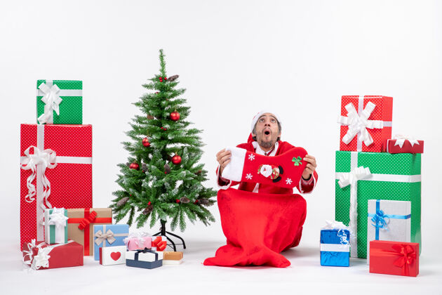 装饰圣诞老人坐在地上 在礼物和白色背景上装饰圣诞树旁展示圣诞袜子惊讶坐着圣诞