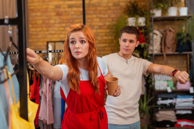 女人购物狂情绪激动的年轻女孩在商店里靠着新衣服 她愤怒的男朋友试图阻止她女性商品男人