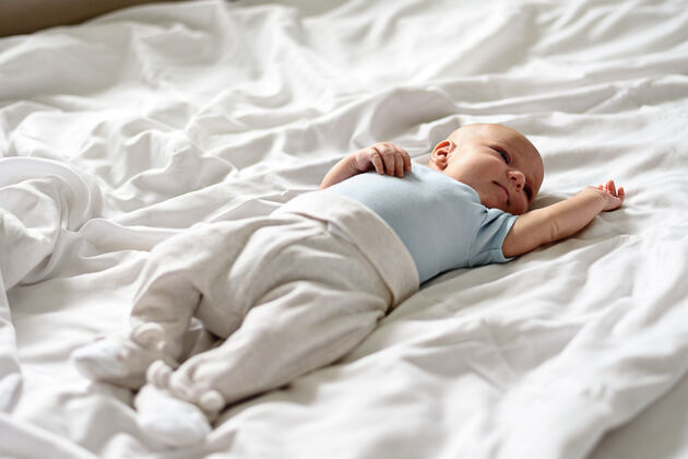 休息穿着蓝色紧身衣和白色裤子的婴儿睁着眼睛躺在白色的床上婴儿小新生儿