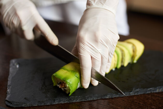 传统制作卷寿司过程的特写镜头手戴手套在黑石板上切卷鳄梨切主人
