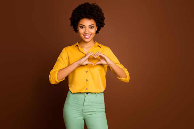 套头衫美丽的深色皮肤波浪女士抱着手臂手指在心脏形状象征心脏健康保健概念穿黄色衬衫绿色裤子孤立棕色的照片喜欢美式混血