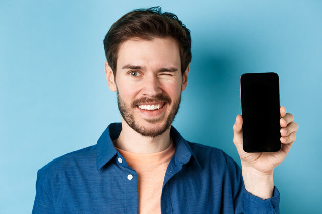 网络特写镜头中英俊的年轻人微笑着 眨着眼睛 展示着空荡荡的手机屏幕 穿着休闲服站在蓝色的背景上交谈在线移动