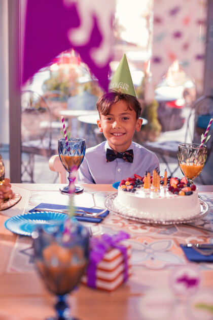 装饰品放松的男孩积极放松的男孩戴着派对帽 微笑着坐在桌子前 面前放着大蛋糕咖啡馆心情童年