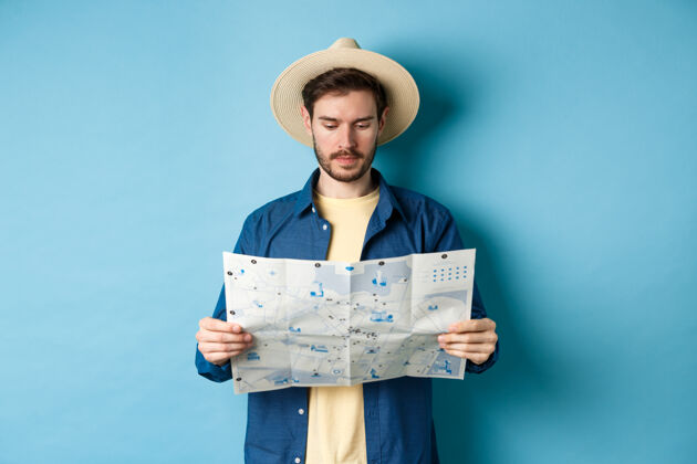 帅气帅气的游客戴着夏帽看地图 寻找度假观光 站在蓝色的背景上男脸男