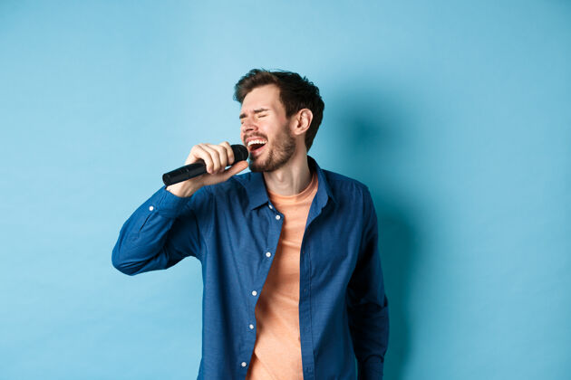 休闲无忧无虑的家伙用麦克风唱卡拉OK 表演一首歌 站在蓝色的背景上复制空间男生肖像朋友