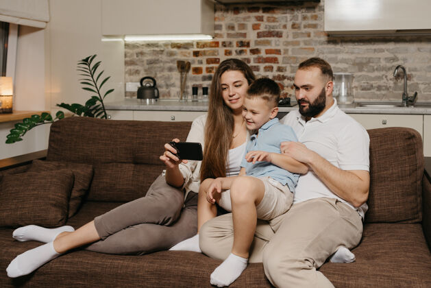 手机一个爸爸 一个儿子和一个年轻的妈妈在沙发上用智能手机看视频童年胡须父亲